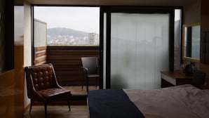Un dormitorio con cama, una silla, un escritorio y vistas a un paisaje urbano desde un balcón.