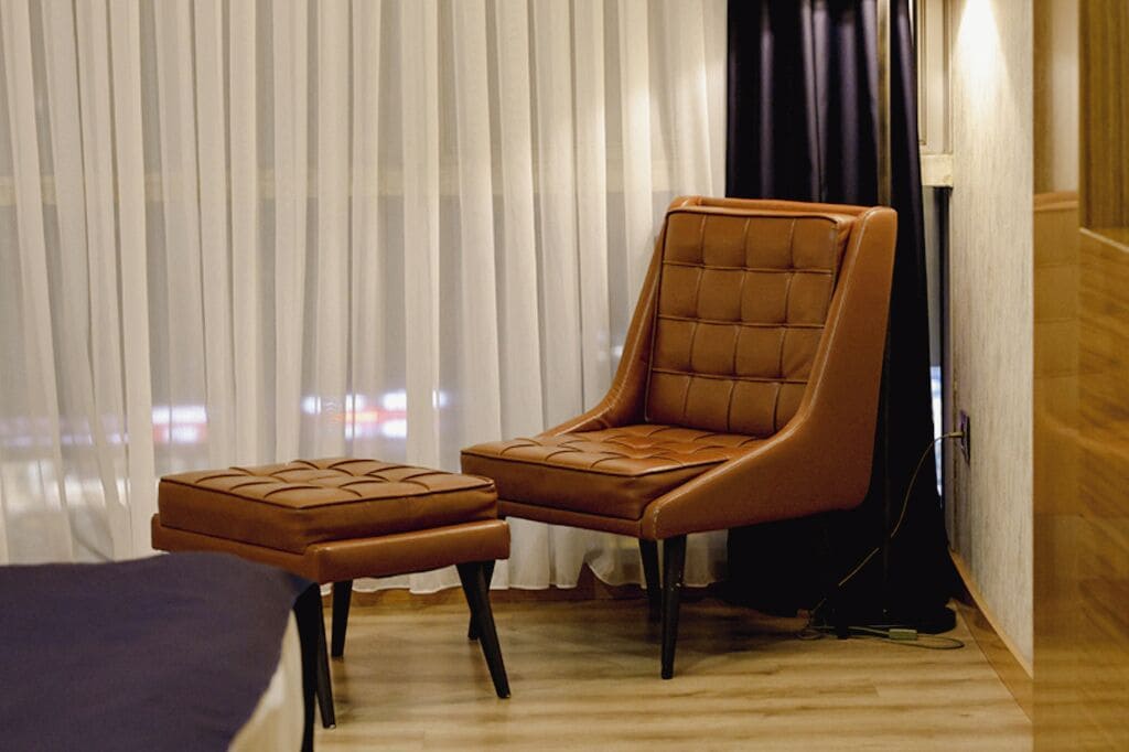 Una habitación moderna con un sillón y reposapiés de cuero marrón, suelo de madera y un ventanal con cortinas translúcidas.