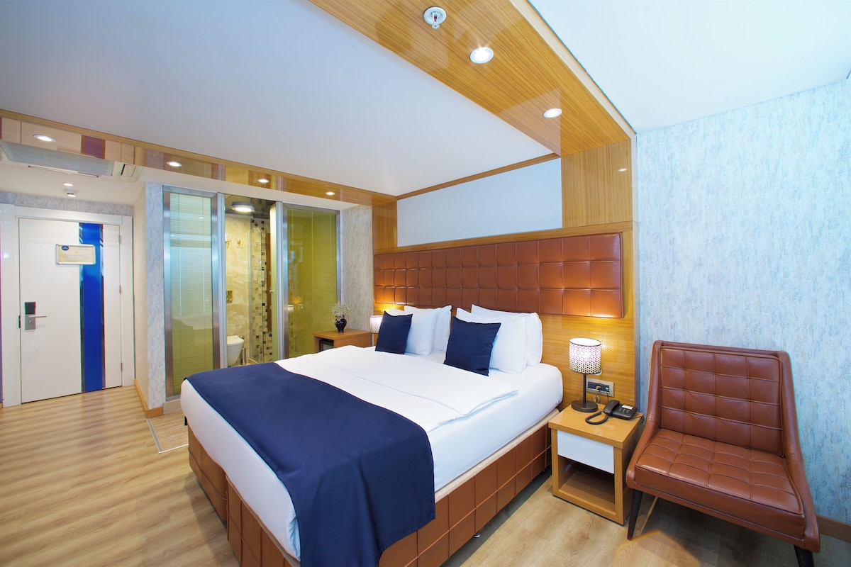 A hotel room with a large bed, a wooden headboard, a desk with a lamp, a chair, and a sliding door leading to a bathroom.