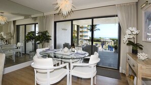 Een moderne eetruimte met een glazen tafel en witte stoelen, een groot raam met uitzicht op de oceaan, en een kroonluchter.