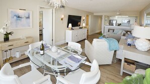 Een moderne woonkamer met een glazen eettafel, witte banken en een keuken op de achtergrond.