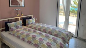 A bedroom with a bed, a bedside lamp, a window with curtains, and a view of the outside. - Villa Aliki (Ierapetra)