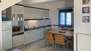 A compact kitchen with white cabinets, a dining table with chairs, and a window with blue curtains. - Villa Aliki (Ierapetra)