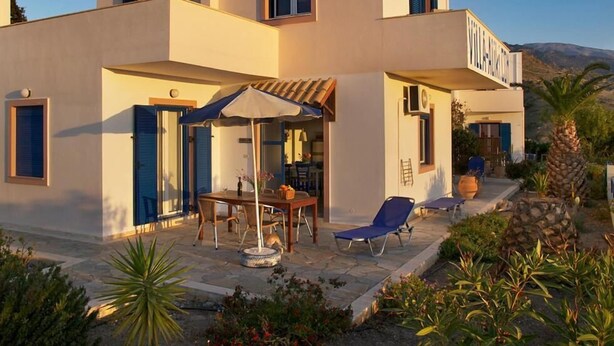 A modern two-story building with a flat roof, a covered outdoor seating area with a table and chairs, and a stone pathway leading to the entrance. - Villa Aliki (Ierapetra)