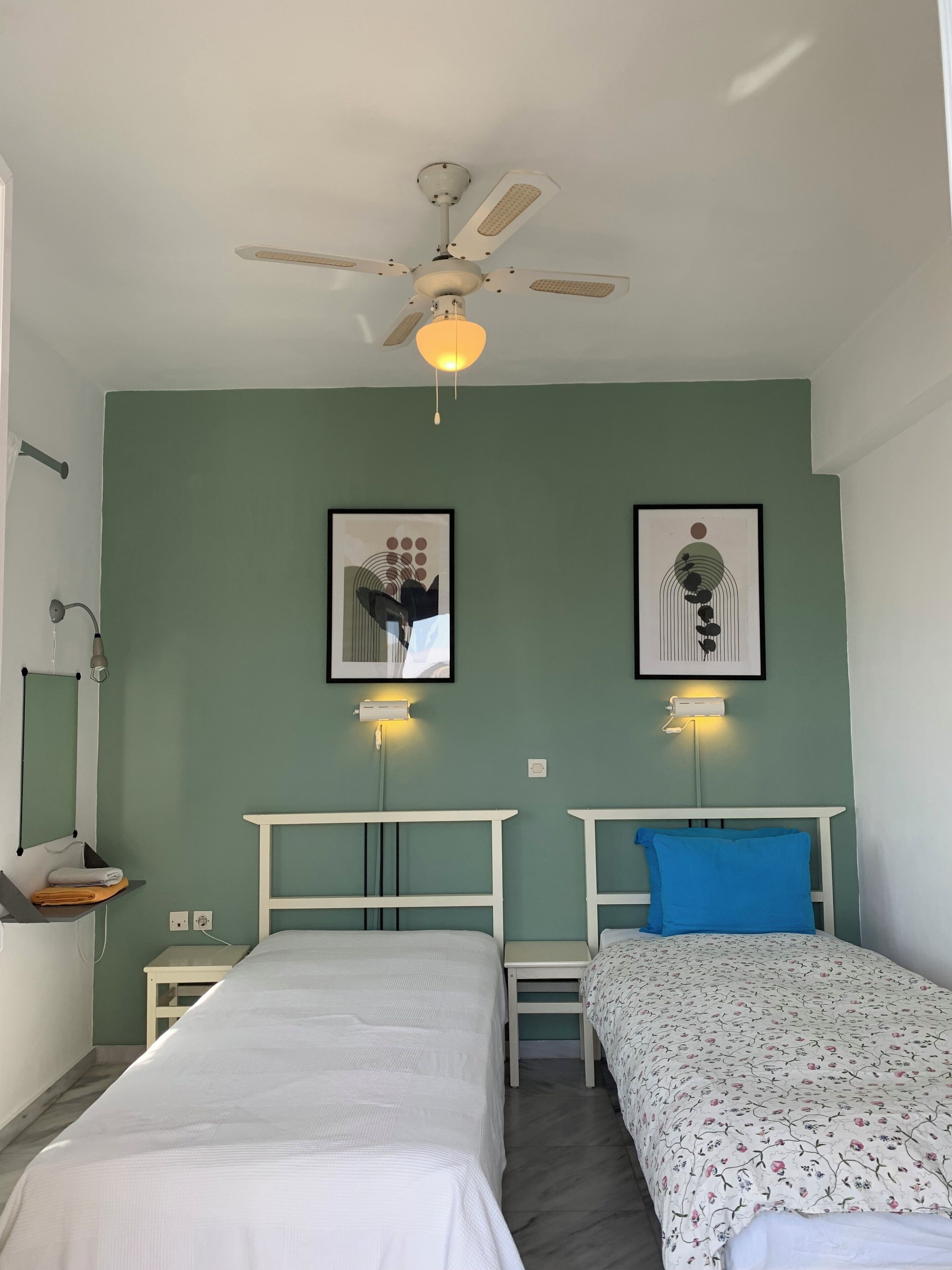 A bedroom with two beds, a ceiling fan, wall-mounted lights, and framed artwork.