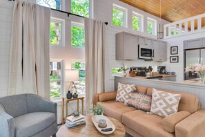 Un salón espacioso con un gran ventanal, un sofá, una mesa de centro y una zona de cocina con microondas y armarios.