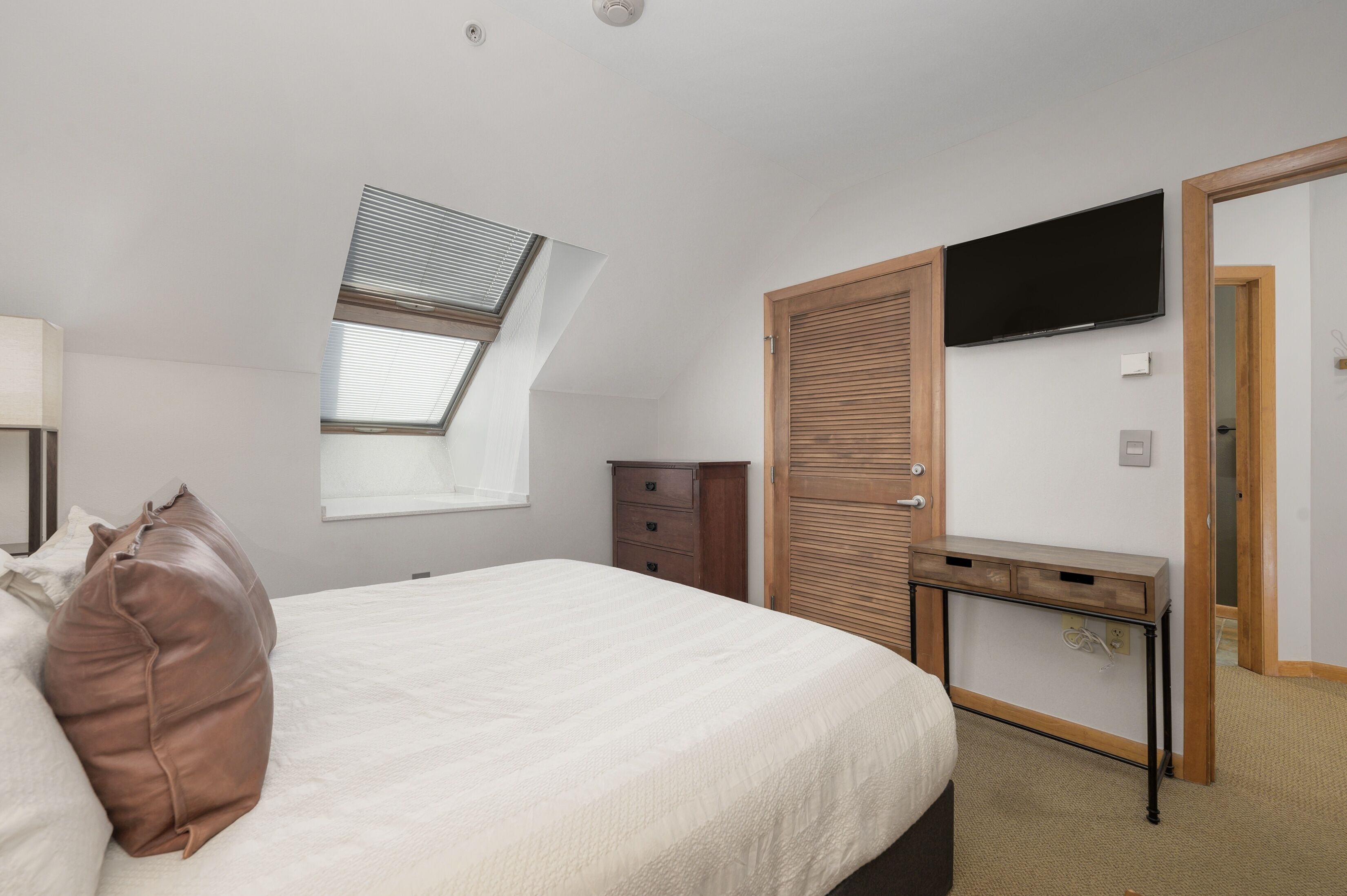 A bedroom with a bed, a desk, a TV, a wardrobe, and a window with blinds.