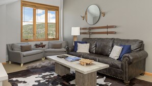 Un salon avec un canapé gris, un canapé en cuir marron, une table basse en bois et une grande fenêtre donnant sur une ville et des montagnes.