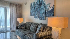A living room with a striped sofa, two lamps, a glass coffee table, and a large abstract painting on the wall. - Ocean Hideaway 610 2 Bedroom Condo by RedAwning (Ocean City)