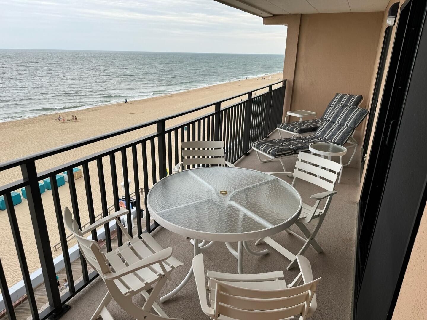 A balcony with a glass table and white chairs, overlooking a beach with people and a calm sea.