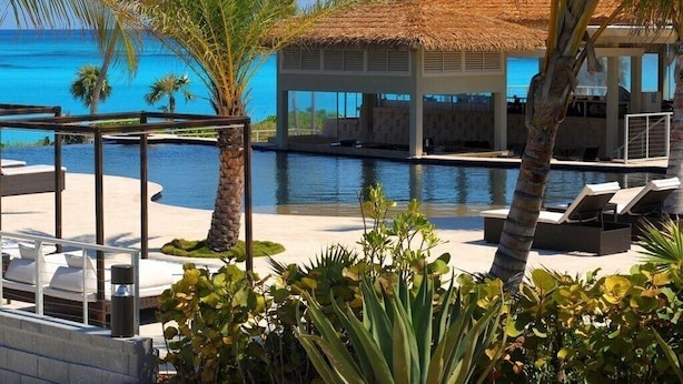 A pool area with lounge chairs, palm trees, and a thatched-roof structure. - Sky Beach Club Rock House 4 Bedroom Home (Governor's Harbour)