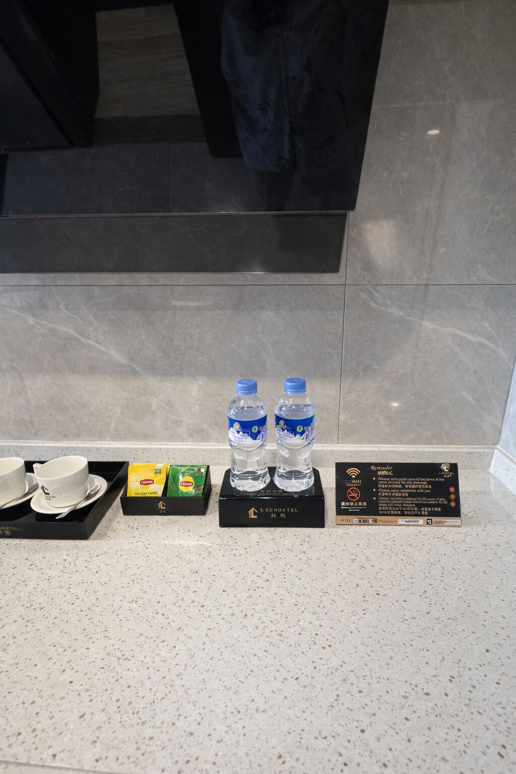 A hotel reception area with a black tray containing two cups and saucers, a box of Lidoine tea bags, and two water bottles on a black stand. A sign with hotel information is also visible.