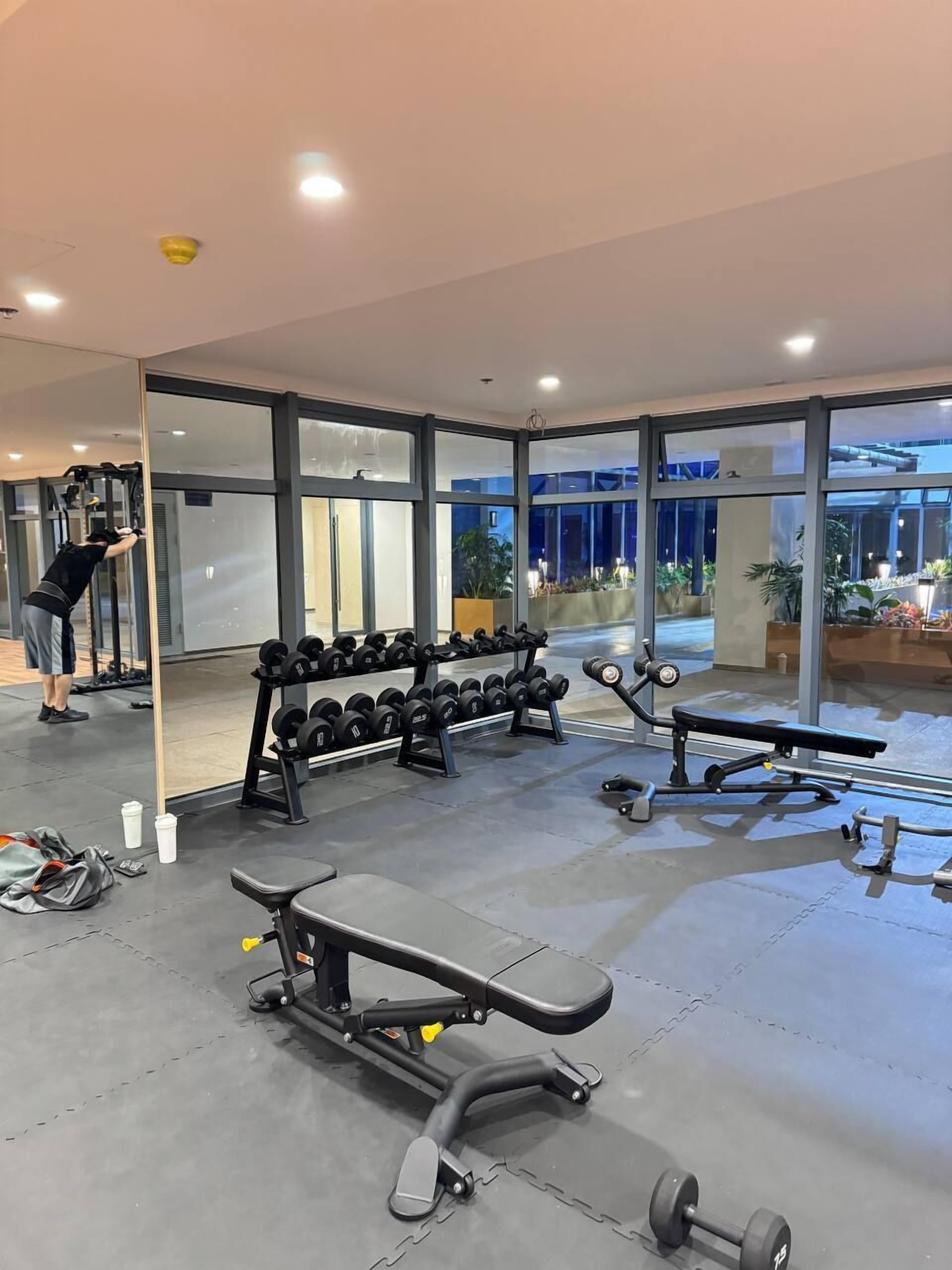 Een moderne sportschool met diverse trainingsapparaten, grote ramen en een persoon die aan het sporten is.