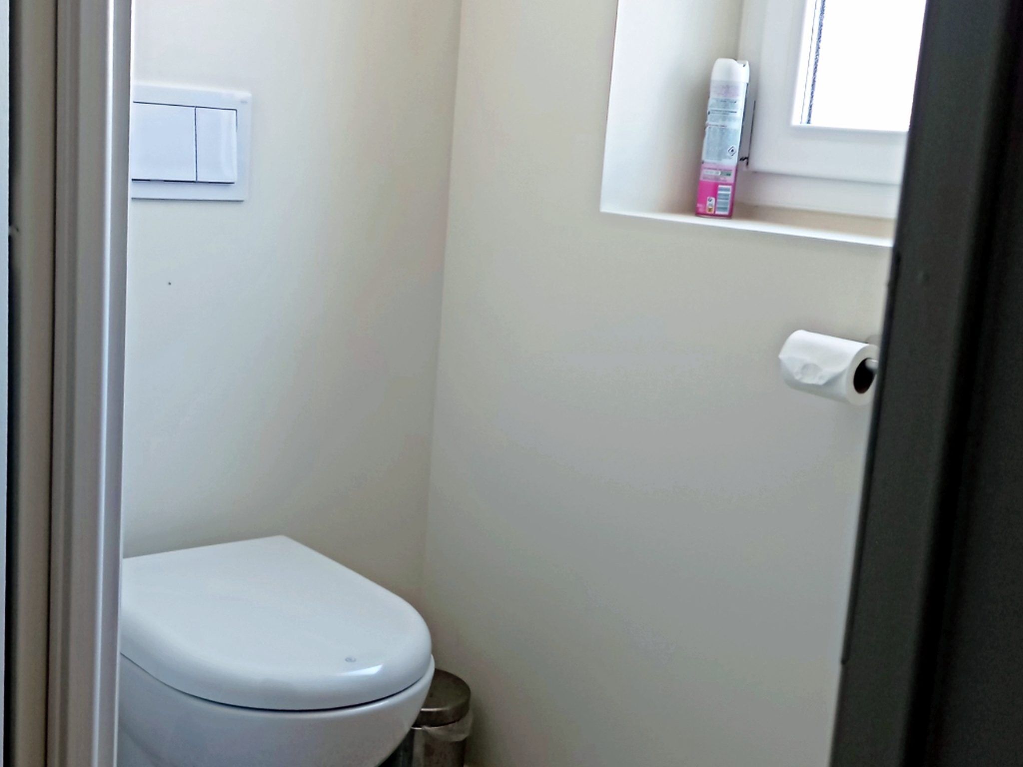 Een kleine, witte badkamer met een toilet, een prullenbak, een toiletrolhouder en een raam met een plank erboven.