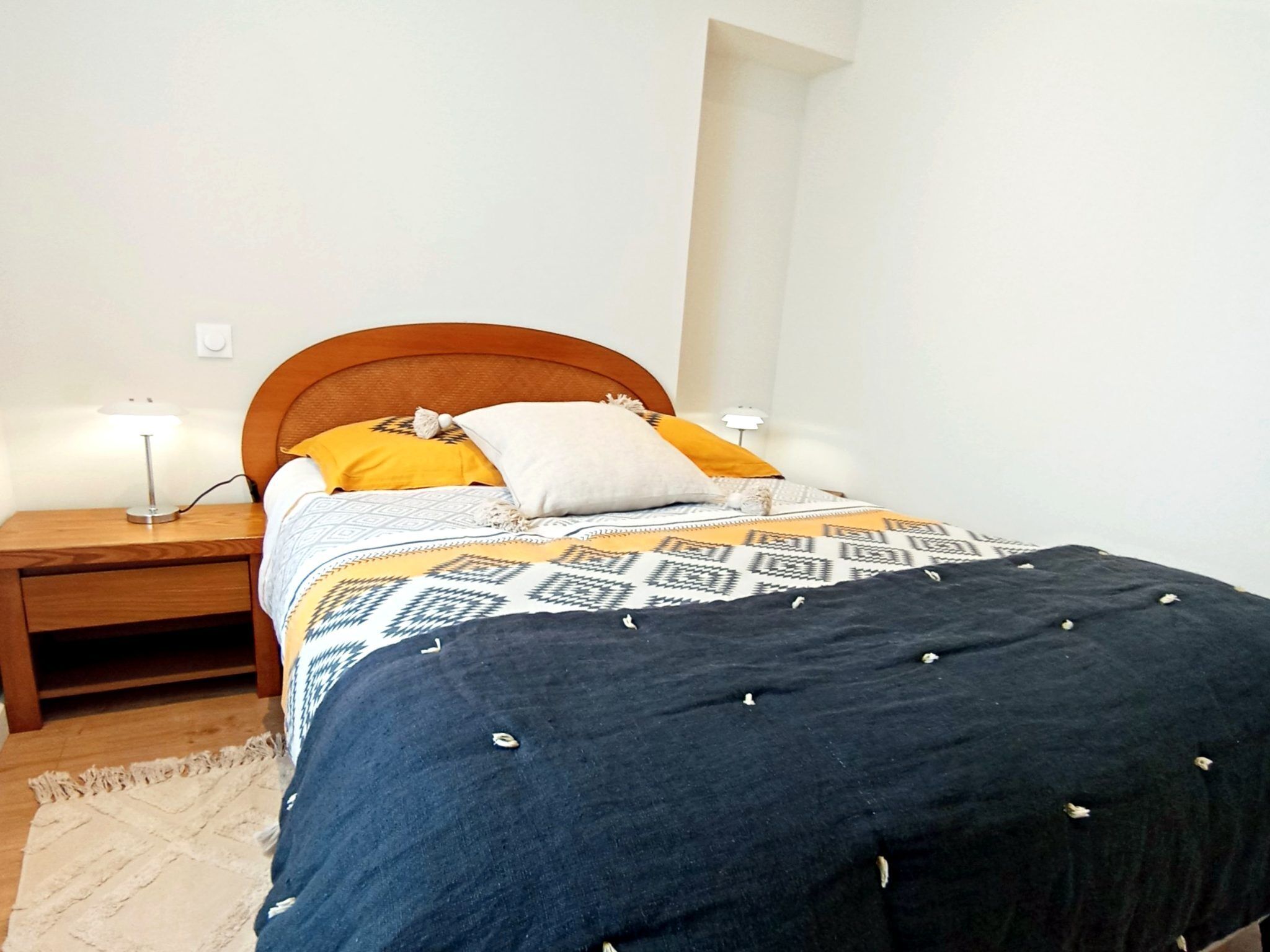 A bed with a patterned bedspread, two bedside lamps, a wooden nightstand, and a rug.
