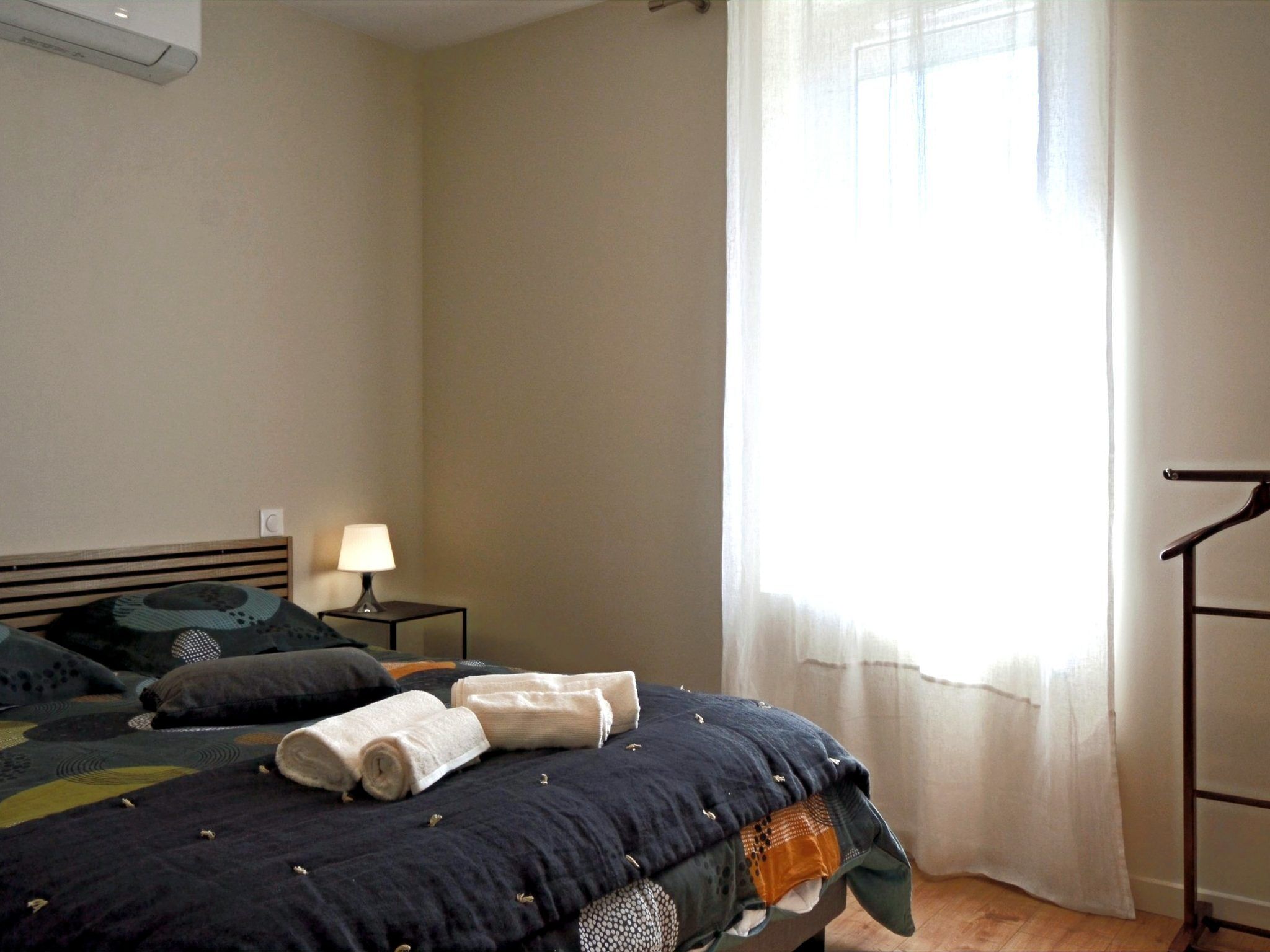A bedroom with a bed, a bedside table with a lamp, a window with curtains, and a wardrobe.