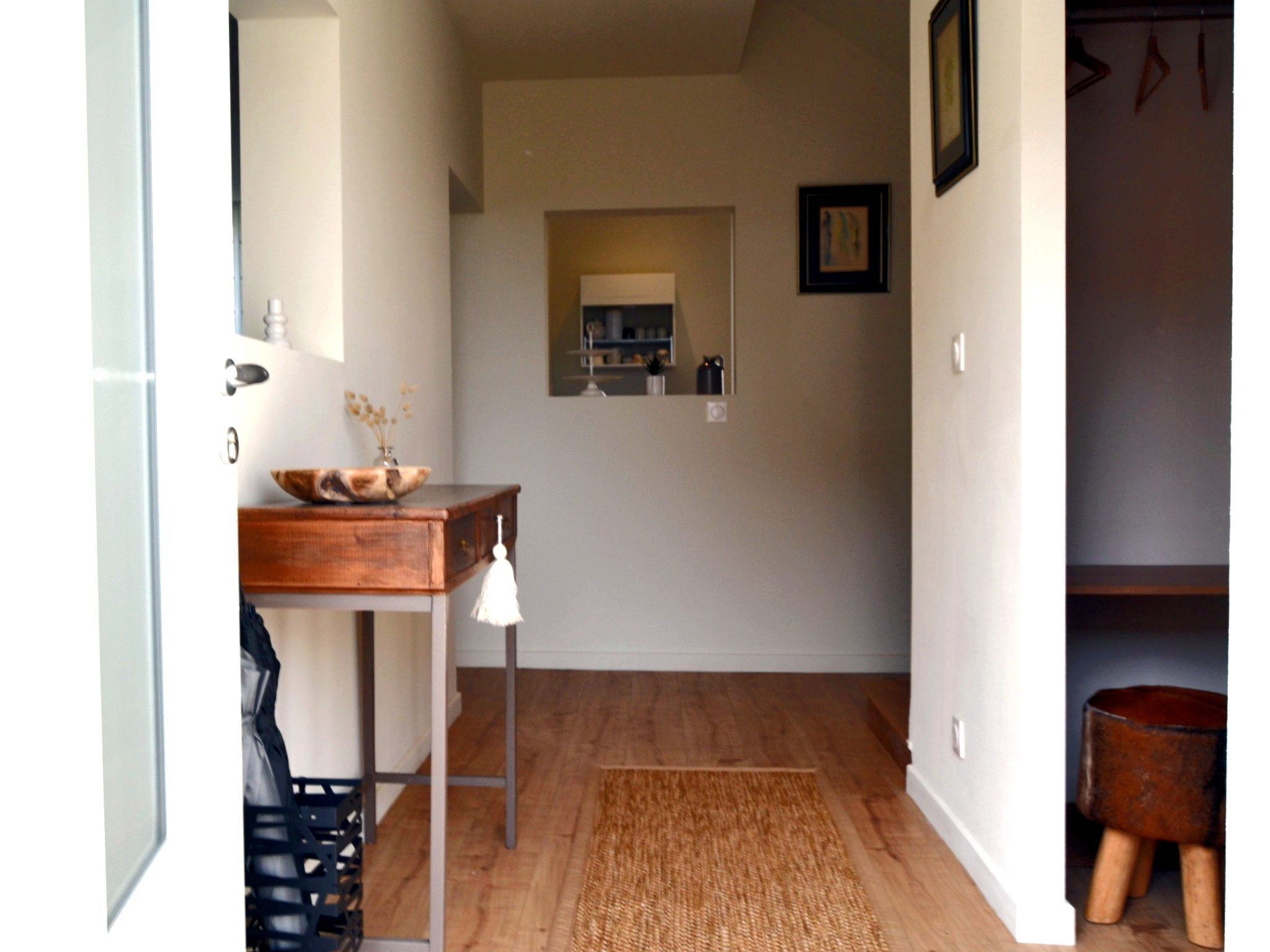A hallway with a wooden floor, a small table with a bowl and vase, a hanging light, and a mirror on the wall.