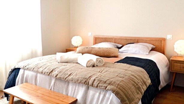 A neatly made bed with rolled towels, a wooden headboard, bedside lamps, and a bench.
