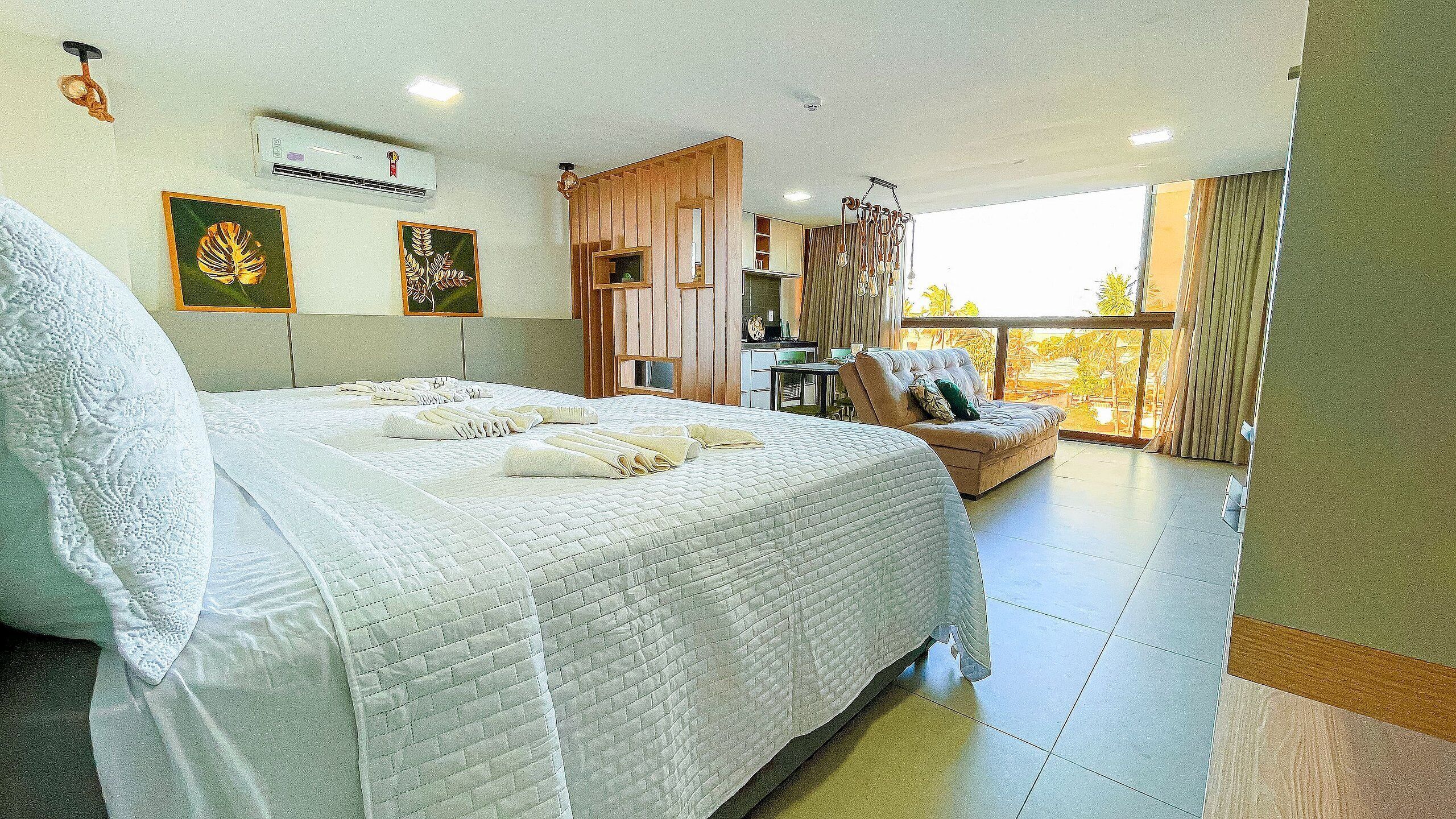 A hotel room with a large bed, a sofa, a dining area, and a balcony with a view.