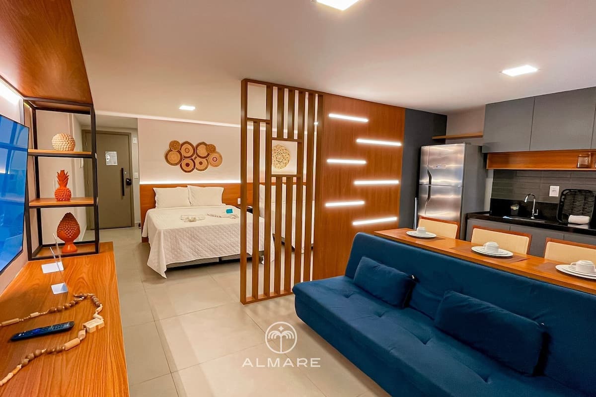 A modern hotel room with a bed, a TV, a desk, a sofa, and a kitchen area.