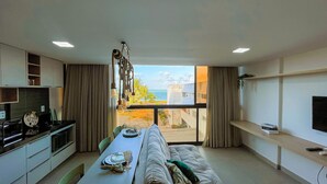 A modern living room with a dining area, large windows with a view of the sea, a kitchen with a stove and cabinets, and a flat-screen TV on the wall.