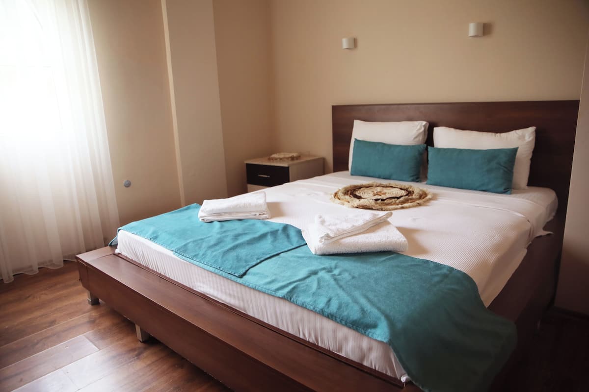 Una cama bien hecha con una colcha color turquesa y ropa de cama blanca, un cabecero de madera y una mesita de noche con una lámpara.