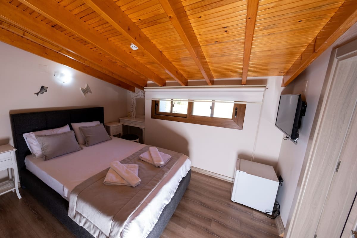 Un dormitorio con techo de madera, una cama con almohadas, una mesita de noche, un aparador, una ventana pequeña y un televisor.