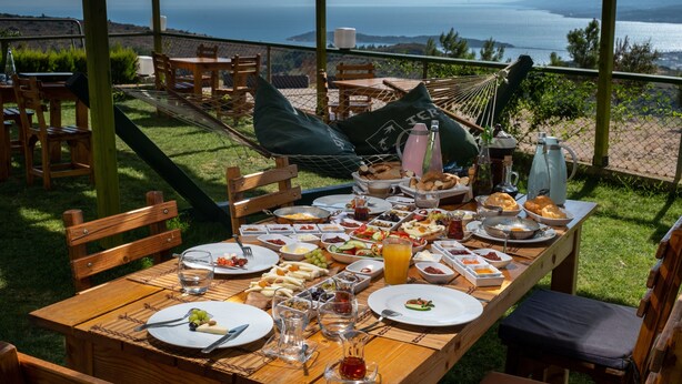 Une table en bois, dressée avec des aliments pour le petit-déjeuner, entourée de chaises et d’un hamac, et offrant une vue panoramique.