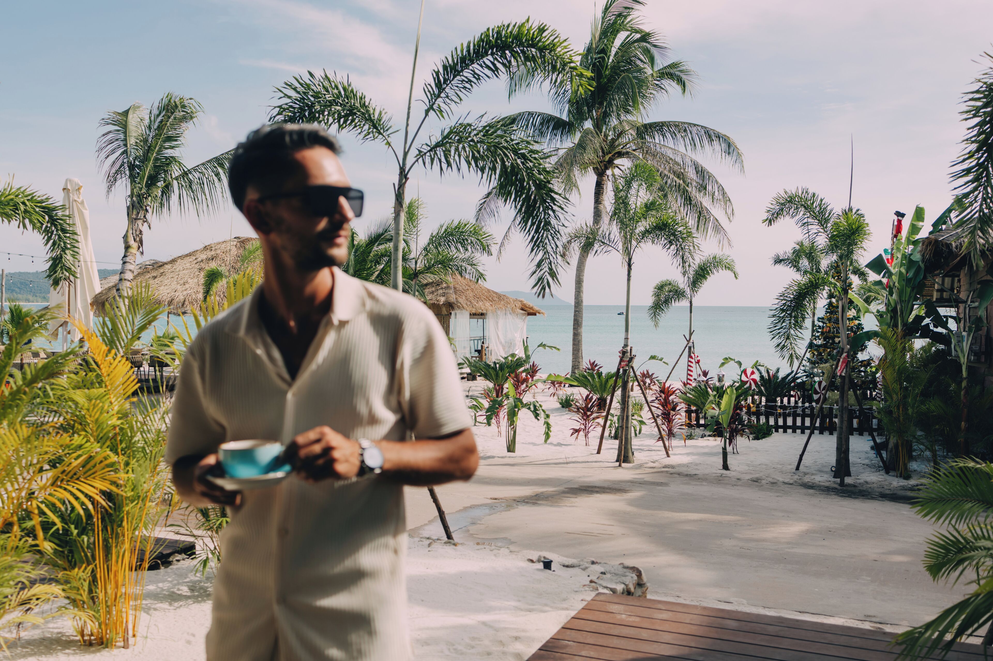 Un homme tenant une tasse, debout sur une terrasse en bois, avec des palmiers et une plage en arrière-plan.