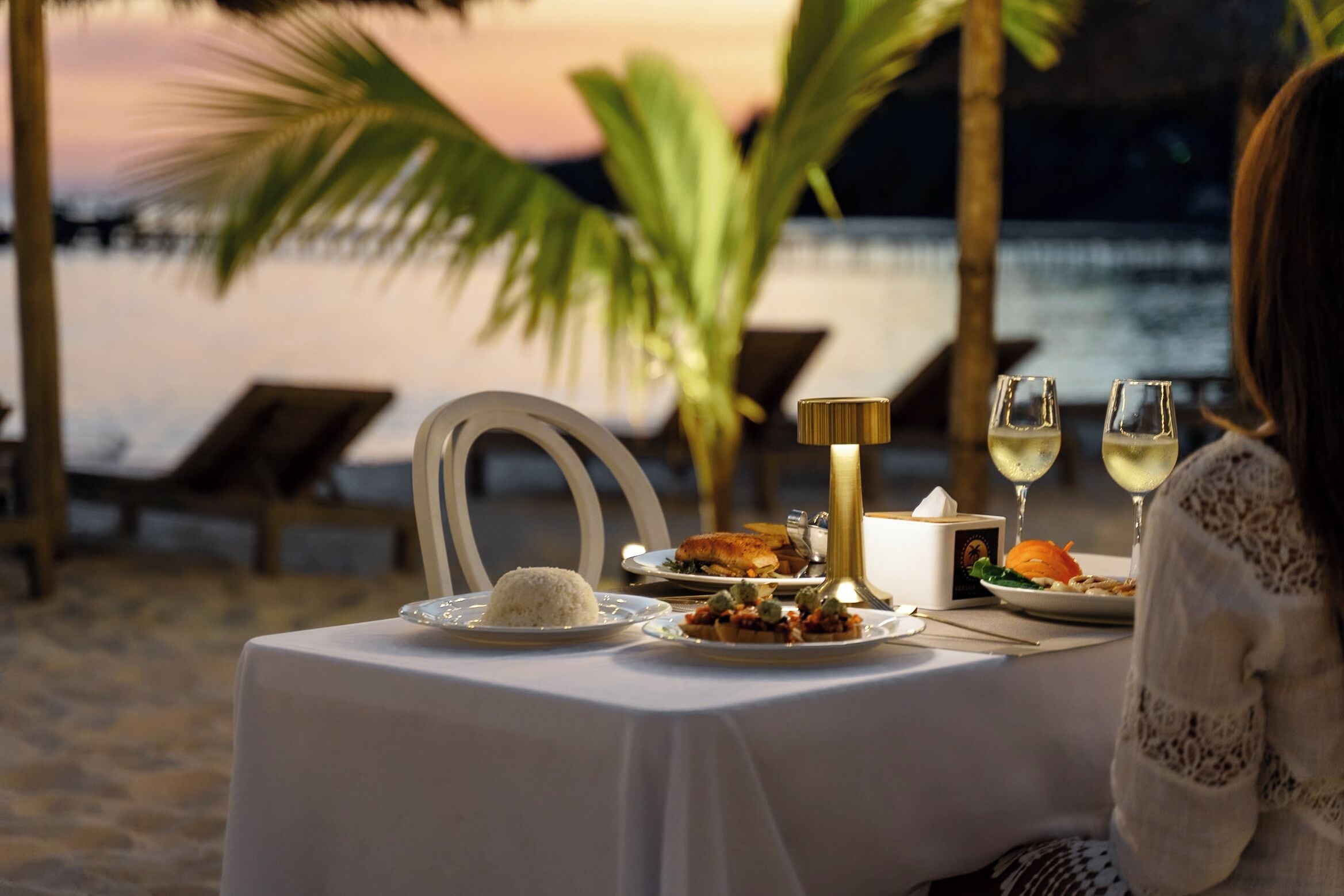 A table set for two with a white tablecloth, plates of food, and two glasses of white wine, overlooking a beach at sunset.