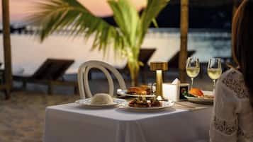 Mesa para dos con mantel blanco, platos de comida y dos copas de vino blanco, con vista a una playa al atardecer.