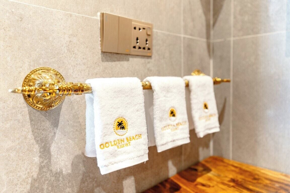 A bathroom with a towel rack holding three white towels with a logo, a gold-colored towel warmer, and a light switch on the wall.
