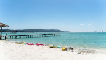 Een strand met een pier die zich uitstrekt over het water, kajaks rijend op het zand, en een constructie met rieten dak.