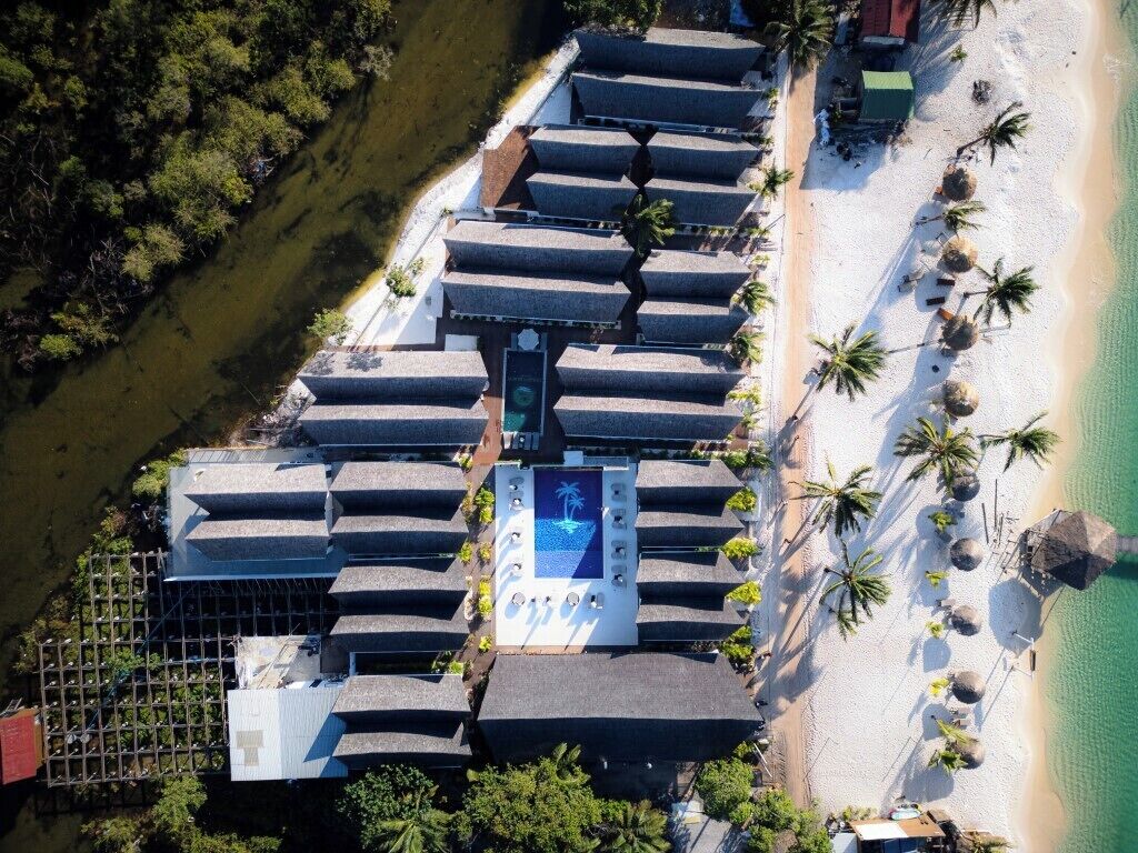 An aerial view of a resort with multiple bungalow-style rooms arranged in a row along a beachfront, a central pool area, and palm trees lining the beach.
