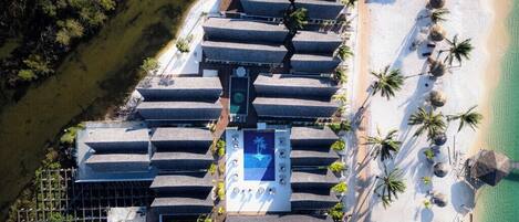 An aerial view of a resort with multiple bungalow-style rooms arranged in a row along a beachfront, a central pool area, and palm trees lining the beach.