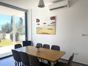 A dining area with a wooden table and six chairs, a potted plant, and a beach-themed painting on the wall. - DPS03 - A Luxury Villa Nissi Pearl - An ultimate stay in Ayia Napa!  (Ayia Napa)