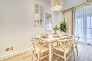 Un coin repas avec une table blanche dressée pour quatre personnes, entourée de chaises en osier, d’un luminaire suspendu et d’une plante en pot.