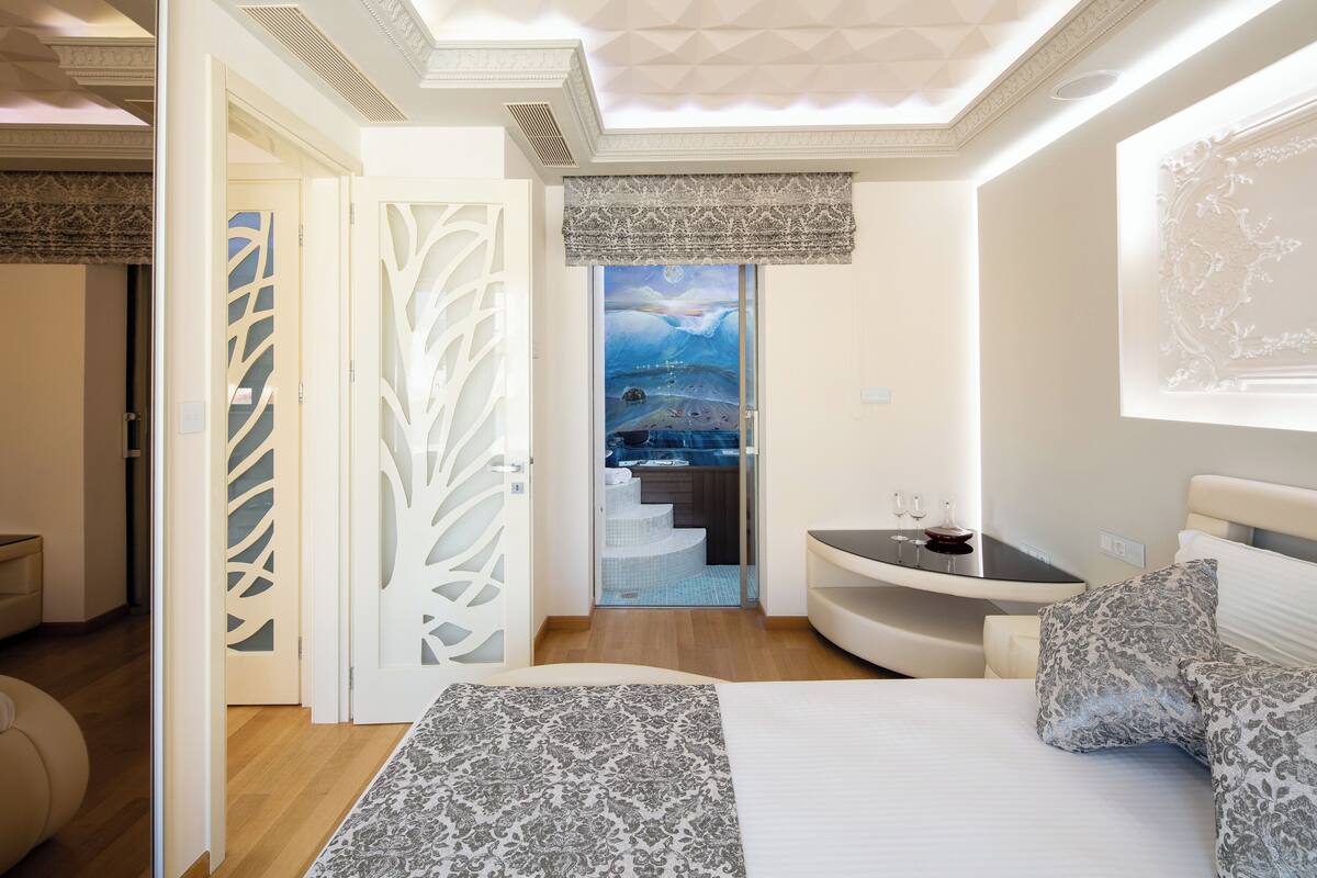 A modern bedroom with a large bed, a small table with glasses, and a view of a kitchen and stairs.
