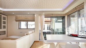 A modern hotel room with a white sofa, a dining table, and a large window with curtains. - DeMaNi President Suites (Sitia)