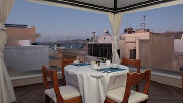 Un coin repas avec une table dressée pour deux personnes, donnant sur un paysage urbain avec des bâtiments et une étendue d’eau.