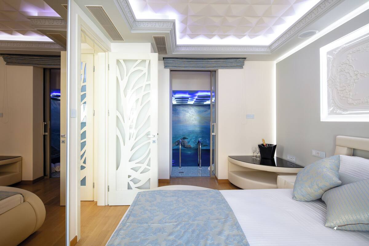 A modern hotel room with a large bed, a desk with a chair, a small seating area, and a view of a pool through a glass door.