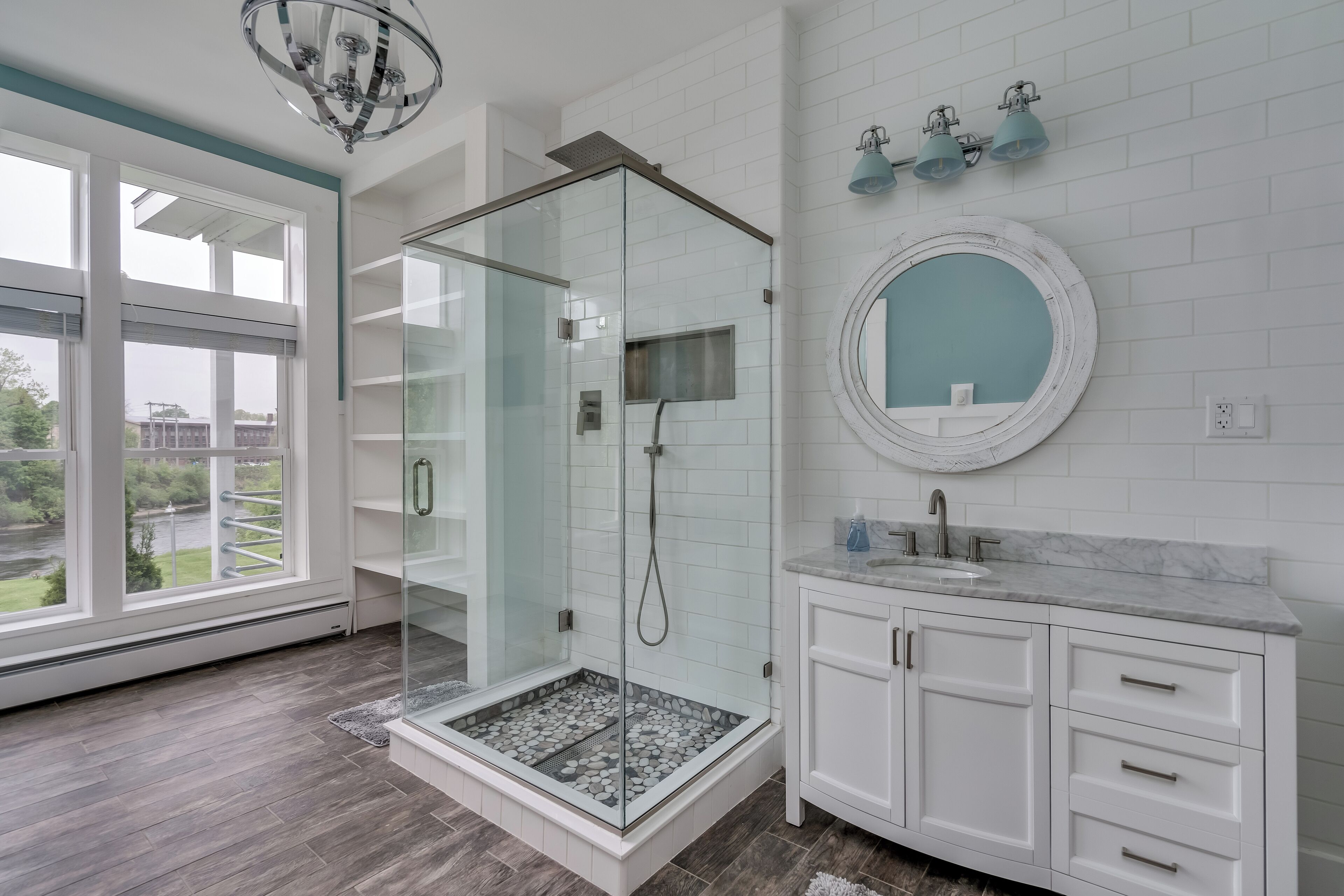 A modern bathroom with a glass-enclosed shower, white cabinetry, a marble countertop, and a large window offering a view of the outdoors.