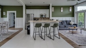 Une cuisine moderne avec des meubles blancs, un îlot central doté de tabourets de bar, et un canapé gris dans un salon lumineux avec de grandes fenêtres.
