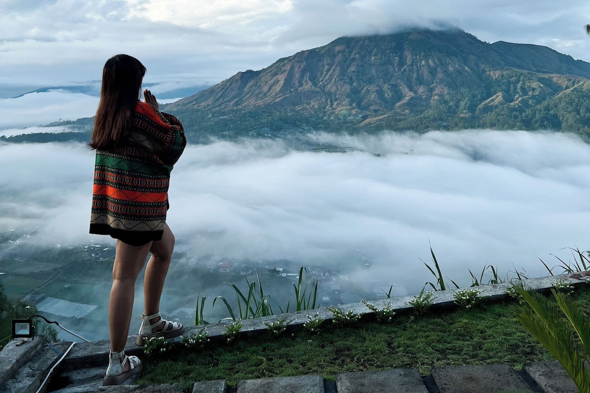 Una persona está sobre una plataforma de piedra contemplando un paisaje montañoso con nubes abajo.