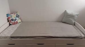 Ein kleines Zimmer mit einem Einzelbett, einer schlichten Matratze, einem farbenfrohen Kissen und einem schlichten Kissen.