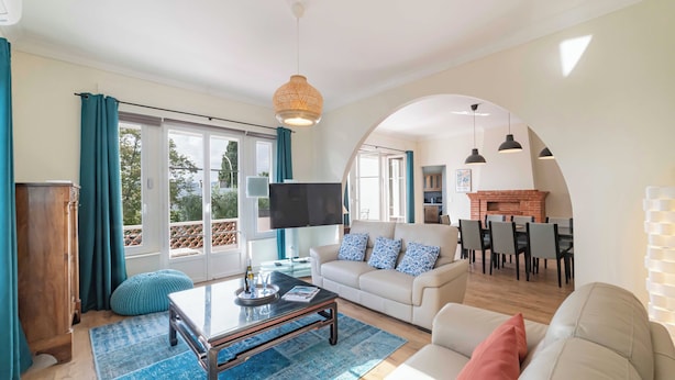 A spacious living room with a sofa, coffee table, and TV. There is a dining area with chairs and a fireplace. Large windows offer a view of the outdoors. - Haut de Villa 6 Personnes Juan les Pins Résidentiel Proche Plage (Antibes)