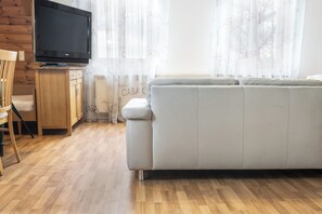 A living room with a grey sofa, wooden flooring, a television on a wooden stand, and a window with curtains.