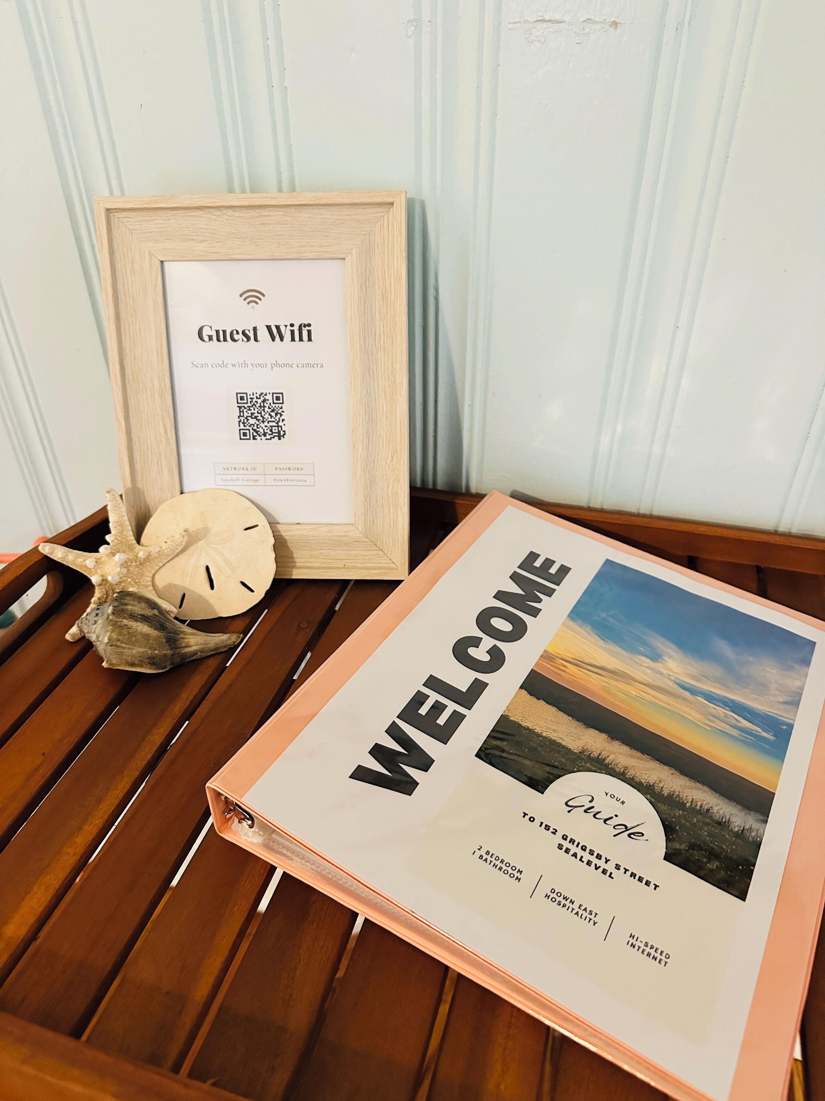 A welcome sign with a scenic view, a framed guest WiFi sign, and seashells on a wooden table.