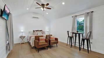 Ein Zimmer mit einem Bett, zwei Sesseln, einem Tisch mit Stühlen, einem Deckenventilator und einem Fenster mit Vorhängen.
