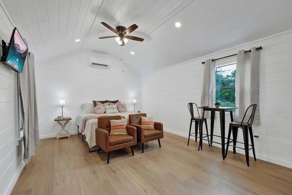 A room with a bed, two armchairs, a table with chairs, a ceiling fan, and a window with curtains.
